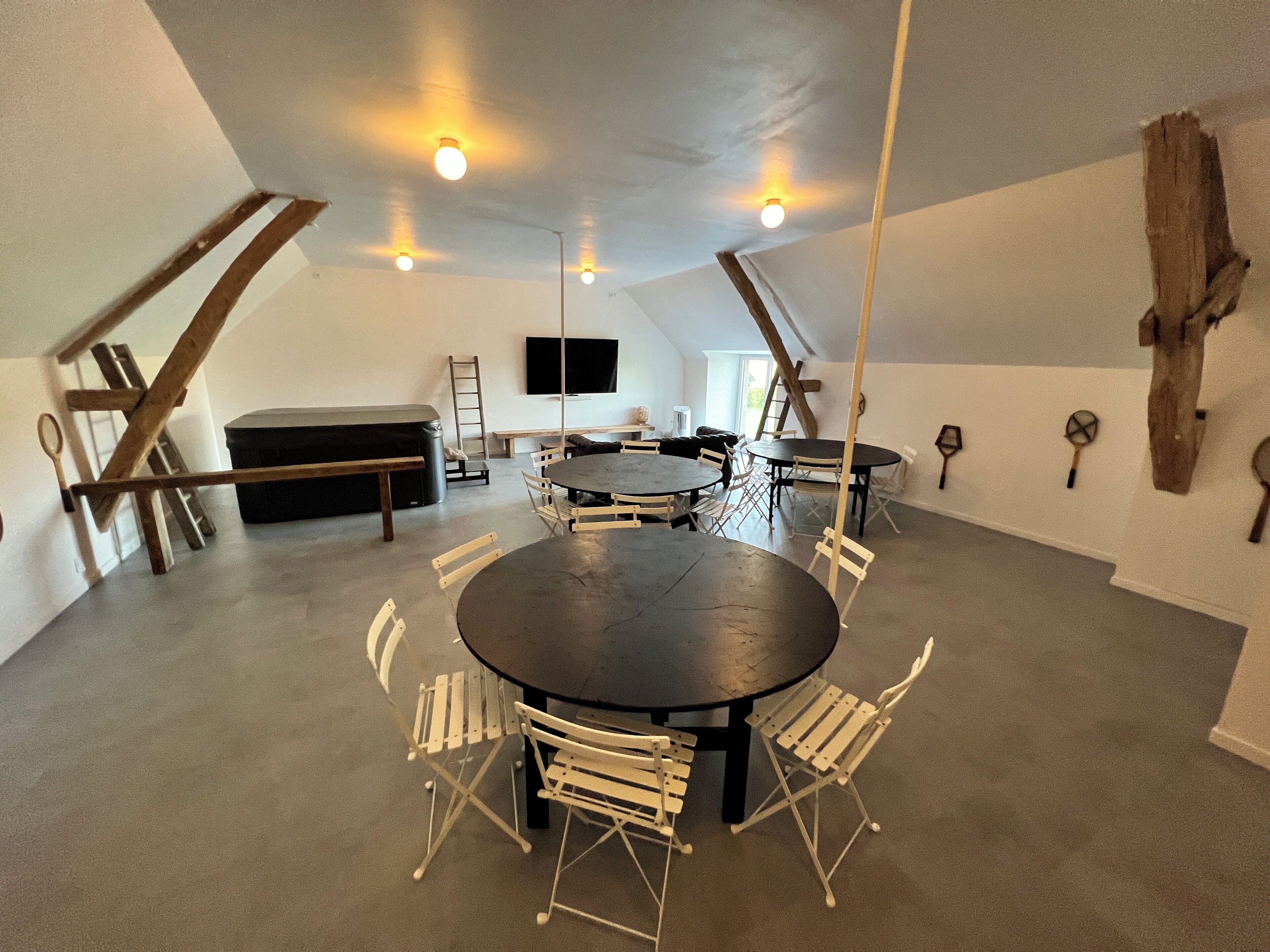 A spacious room with a large round table and white folding chairs, a sofa, and a television mounted on the wall.