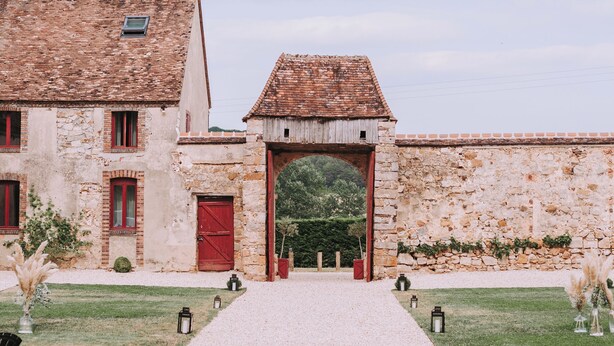 Un bâtiment avec un chemin de gravier menant à une porte rouge, entouré de murs de pierre et de verdure.