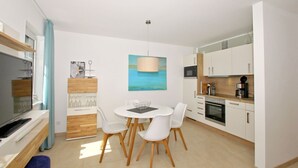 A modern kitchen with white cabinets, a dining area with a round table and white chairs, a TV, and a painting on the wall. - Beachfront apartment with terrace, use of swimming pool and sauna - Rex Rugia H-1010 - (Mönchgut)