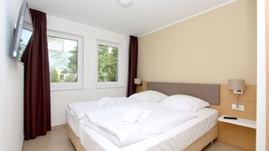 Una habitación de hotel con una cama grande, dos ventanas, un televisor montado en la pared y una mesita de noche con lámpara.