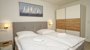 Een net opgemaakt bed met witte lakens, een grijs gestoffeerd hoofdbord en een houten paneel aan de muur.