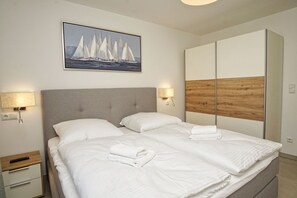 Ein ordentlich bezogenes Bett mit weißen Leinentüchern, einem grau gepolsterten Kopfteil und einer Holzpaneele verkleideten Wand.