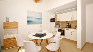 Una cocina compacta con armarios blancos, microondas y cafetera. Hay una zona de comedor con una mesa redonda y sillas blancas. Un cuadro cuelga en la pared sobre la mesa.