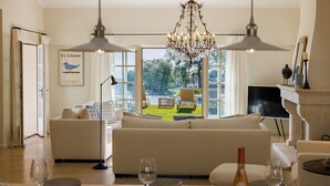 A spacious living room with a large sofa, dining table set for a meal, and a view of a garden through large windows. - Mas Bellevue – Heated Pool & Exceptional Sea View (Saint Paul de Vence)