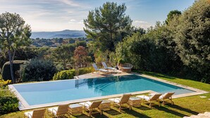 A rectangular swimming pool with lounge chairs and a wicker chair, surrounded by well-maintained grass and trees, with a view of distant hills. - Mas Bellevue – Heated Pool & Exceptional Sea View (Saint Paul de Vence)