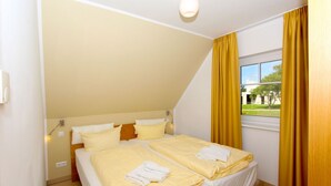 Un dormitorio con dos camas, techo inclinado, ventana con cortinas amarillas y vista a un edificio.