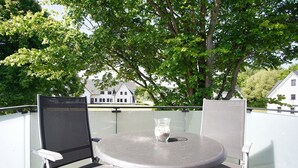 Ein runder Tisch mit zwei Stühlen auf einem Balkon, eine Glasvase mit Blumen und eine Ansicht von Häusern im Hintergrund.