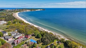 Ovenudblik over et kystområde med en bugtet strand, boligbygninger og et klart, blåt hav.