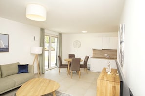 A modern living room with a sofa, coffee table, dining table, and chairs. There is a kitchen area with cabinets and a window. - Beachfront apartment with terrace, use of swimming pool and sauna - Rex Rugia H-1010 - (Mönchgut)