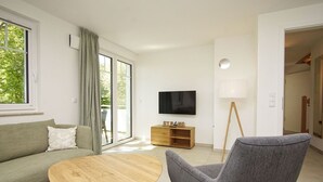 Ein modernes Wohnzimmer mit einer Couch, einem runden Holztisch, einem grauen Sessel, einem an der Wand befestigten Fernseher und einer Holzkommode mit dem Schriftzug "STRAND".