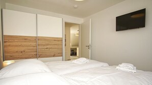 Quarto com duas camas, cabeceira de madeira, televisão na parede e uma porta que dá para outro espaço.