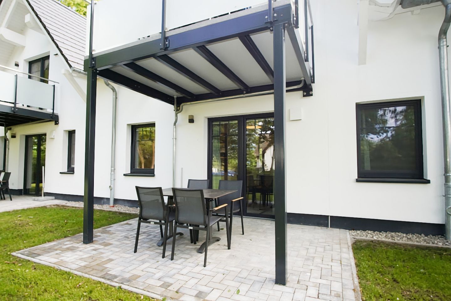 Une terrasse extérieure avec une table et des chaises couvertes, un bâtiment blanc doté de grandes fenêtres et une zone dallée.