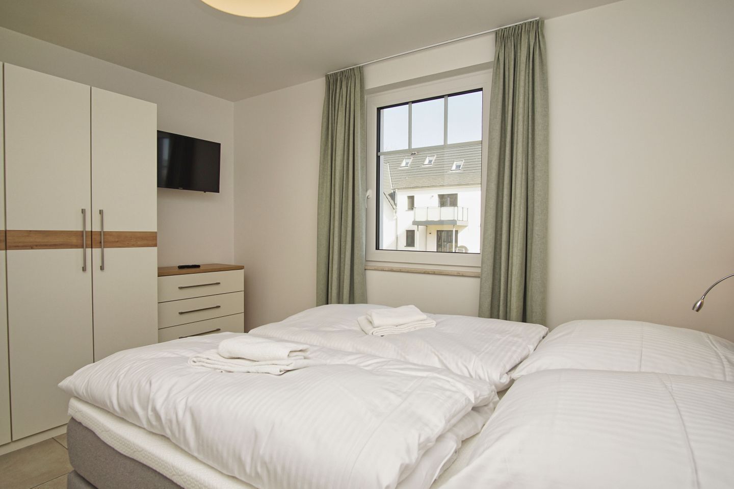 A bedroom with a bed, a TV mounted on the wall, a window with curtains, and a wardrobe.