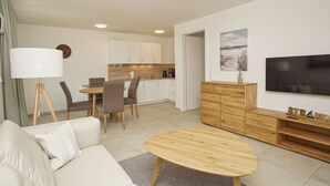 Een moderne woonkamer met een bank, een salontafel, een eethoek met stoelen, een keuken op de achtergrond en een televisie aan de muur.