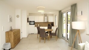 Une cuisine moderne avec des meubles blancs, un coin repas avec une table et des chaises en bois, et une grande fenêtre avec des rideaux.