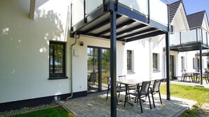 Une terrasse extérieure moderne, dotée d’une table et de chaises, d’un espace salon abrité et d’une porte vitrée donnant sur un jardin.