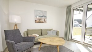 A modern living room with a grey armchair, a green sofa, a wooden coffee table, a lamp, and a painting of seagulls on a pier. - Beachfront apartment with terrace, use of swimming pool and sauna - Rex Rugia H-1010 - (Mönchgut)