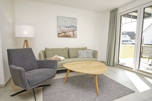 Ein modernes Wohnzimmer mit einem grauen Sessel, einem grünen Sofa, einem Holztisch, einer Lampe und einem Gemälde von Möwen auf einem Steg.
