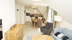 Ett modernt vardagsrum med ett soffbord i trä, en grå fåtölj och en blå soffa. Det finns ett TV-bord i trä och en matplats med ett bord och stolar i trä.