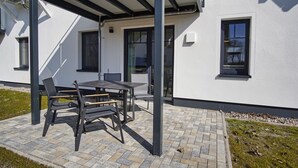 Eine Terrasse im Freien mit Tisch und Stühlen, ein überdachter Sitzbereich und ein Gebäude mit großen Fenstern und einer Tür.