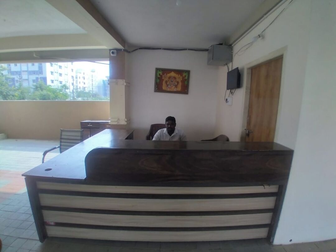 A reception desk with a person seated behind it, a framed picture on the wall, and a window with a view of buildings and trees.