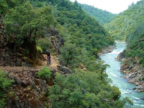 Ciclista de montaña en un sendero pedregoso, con un río al fondo.