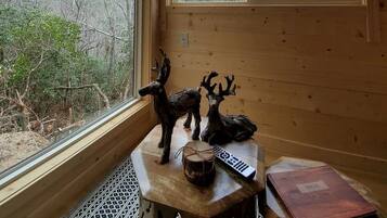 A cabin interior with wooden walls, a mounted television, a window with a view of trees, and a small table with a deer figurine and a book.