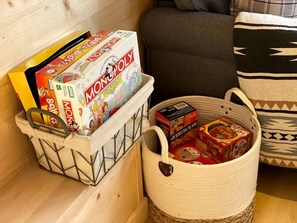 A storage basket with Monopoly Junior and Monopoly games, a woven basket with a Hello Kitty box, and a patterned pillow on a wooden floor. - Stunning Tiny House in Nature Perfect for Luxury Camping in Hiawassee, Georgia (Hiawassee)