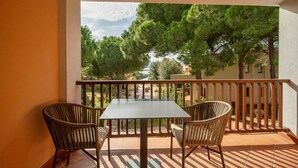 Varanda com mesa e duas cadeiras de vime, com vista para um jardim com árvores e um edifício ao fundo.