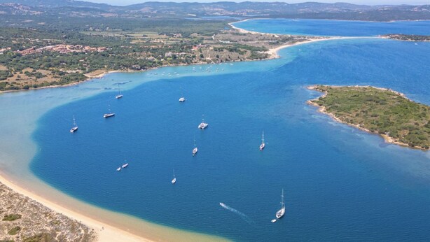 Vista aérea de uma área costeira com águas azul-claras, vários barcos e uma praia de areia.