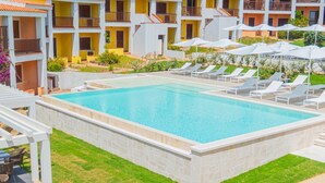 Piscina com espreguiçadeiras e guarda-sóis, rodeada por um gramado bem cuidado e um edifício com varandas.