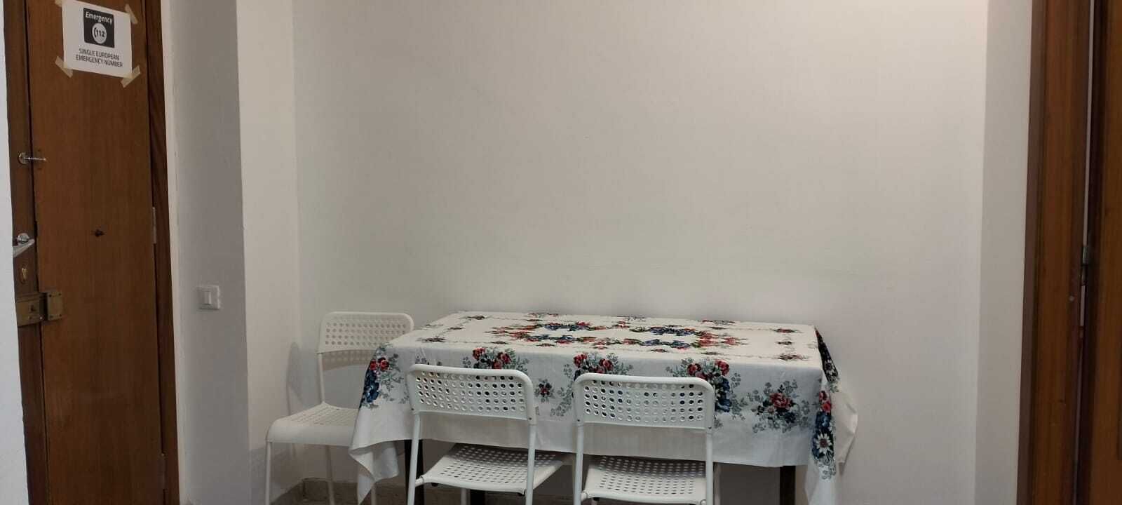 A small room with a table and two chairs, a wooden door with an emergency number sign, and a plain white wall.