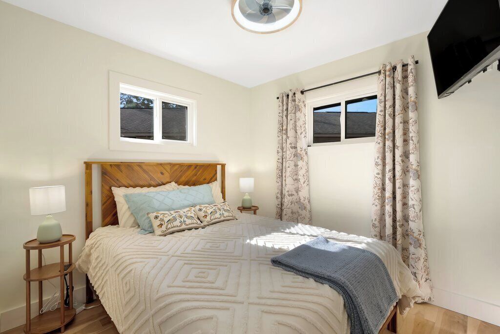 Een slaapkamer met een houten hoofdbord, een bed met beddengoed met een patroon, een nachtkastje met een lamp, een raam met gordijnen, en een flatscreen televisie aan de muur.