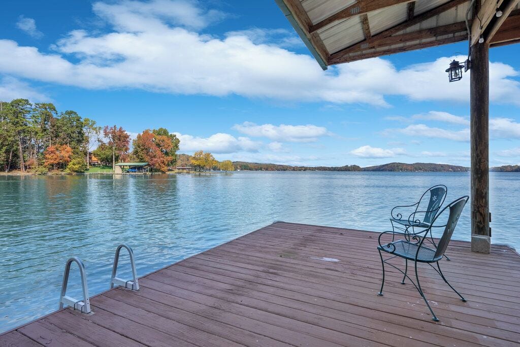 A wooden deck with metal chairs overlooking a calm lake, surrounded by trees and a clear sky.