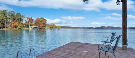 A wooden deck with metal chairs overlooking a calm lake, surrounded by trees and a clear sky.