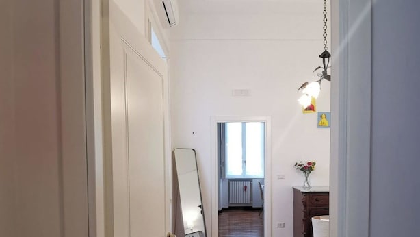 A hallway with a wooden floor, a bed with white bedding, a hanging light fixture, and a door with a number plate. - Comfortable rooms in the heart of Vomero, just a few meters from the subway. (Napoli)