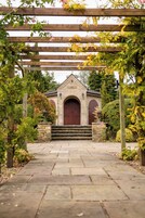 Un edificio de piedra con una entrada de pérgola de madera, una escalera de piedra y un camino pavimentado que conduce al edificio.