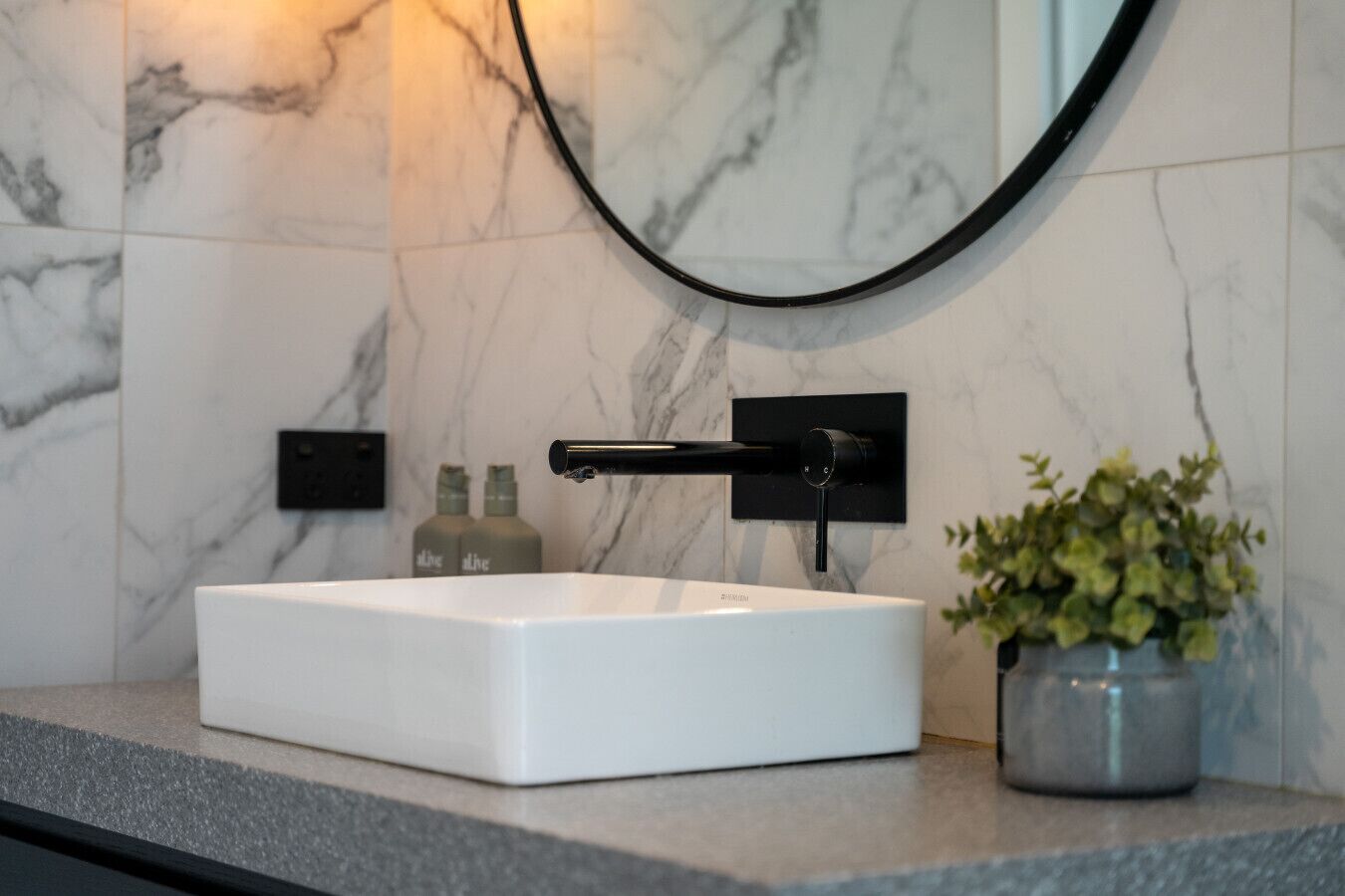 A white rectangular sink with a modern faucet, mounted on a marble wall with a black fixture above it. There are two bottles with labels on the wall to the left of the sink.