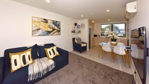 A living room with a sofa, a dining area with a table and chairs, a kitchen in the background, and a large window with a view. - Central 2BR Townhouse Close to Te Kaha Stadium (Christchurch)