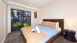 A hotel room with a large bed, a bedside lamp, a view of a garden through a sliding door, and a painting on the wall. - Stylish 2BR Retreat by Te Kaha Stadium (Christchurch)