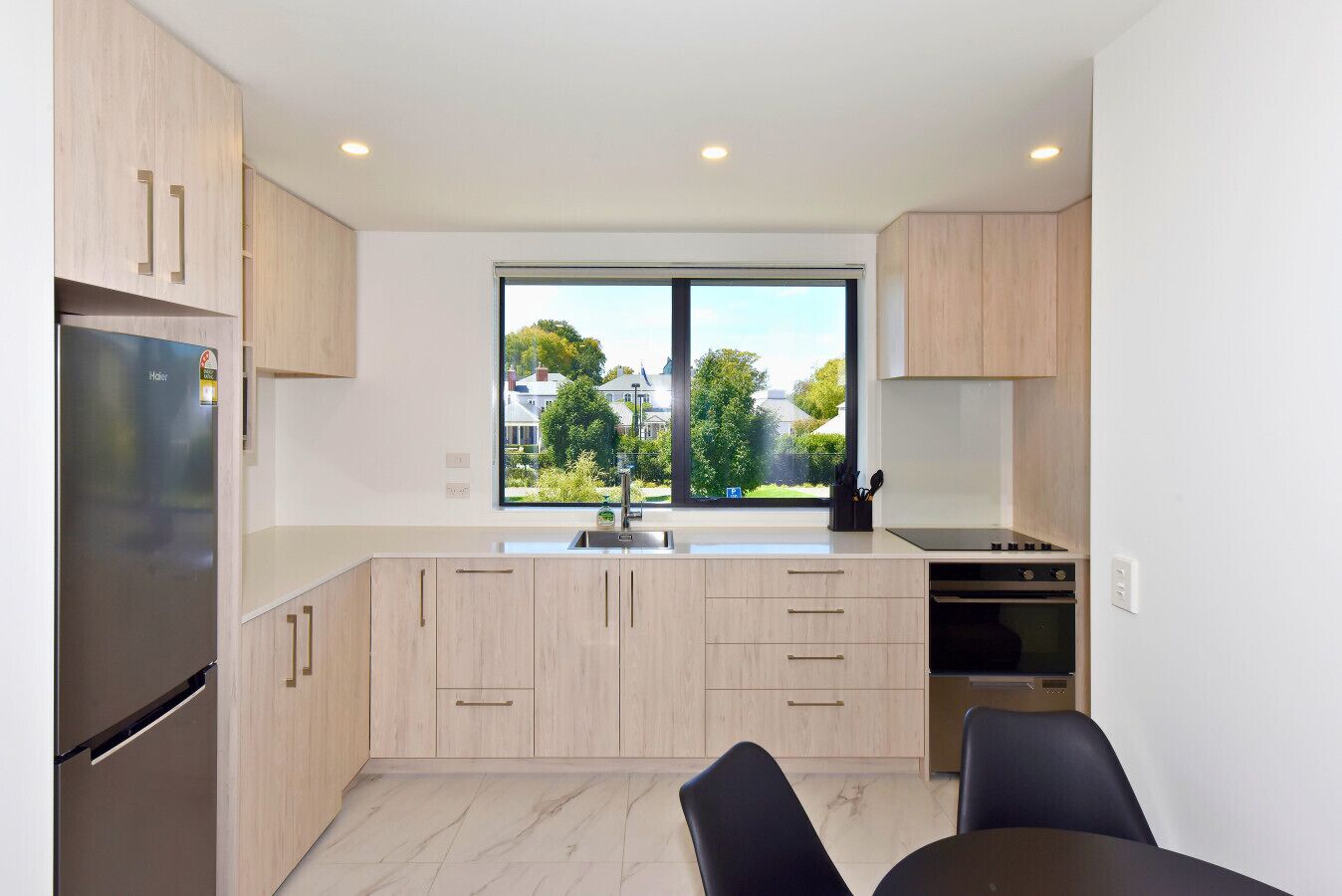 Een moderne keuken met lichte houten kastjes, een roestvrijstalen koelkast, een raam met uitzicht op huizen, en een eethoek met twee stoelen.