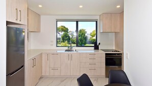 A modern kitchen with light wood cabinets, a stainless steel refrigerator, a window with a view of houses, and a dining area with two chairs. - Stylish 2BR Retreat by Te Kaha Stadium (Christchurch)