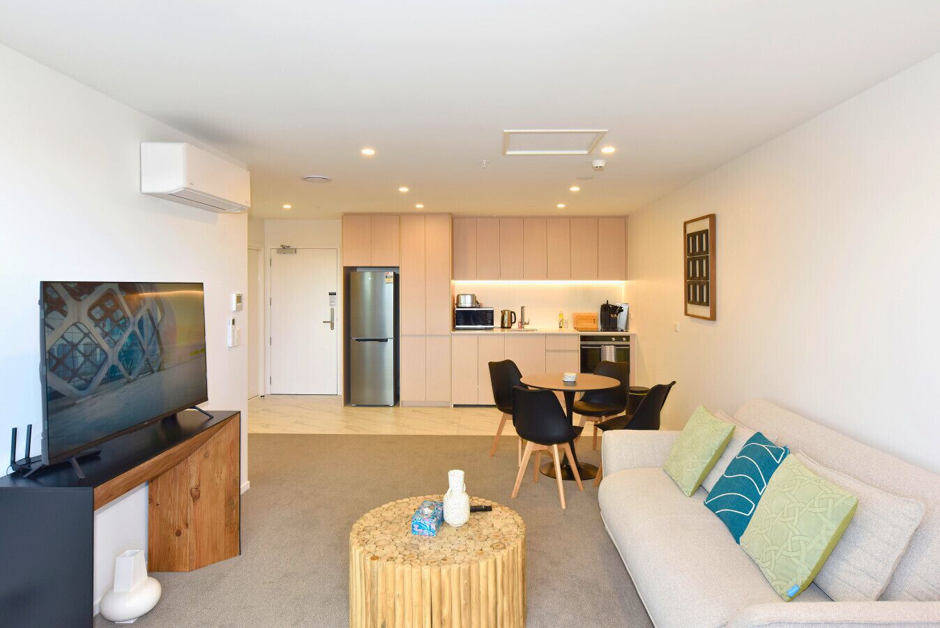 A modern living room with a flat-screen TV, a wooden coffee table, a sofa with patterned cushions, a kitchen area with a refrigerator, microwave, and dining table with chairs.