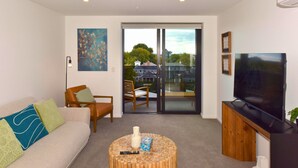 A living room with a sofa, chairs, a coffee table, a TV, and a sliding glass door leading to a balcony. - Fresh 1 Bedroom Apartment Close to Te Kaha Stadium (Christchurch)