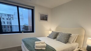 Une chambre moderne avec un grand lit, une table de chevet, une fenêtre donnant sur des immeubles et un petit tableau accroché au mur.