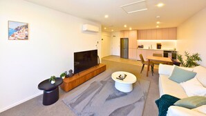 Ein modernes Wohnzimmer mit Flachbildfernseher, Couchtisch und Sofa. Es gibt eine Küchenzeile mit Schränken und einem Kühlschrank.