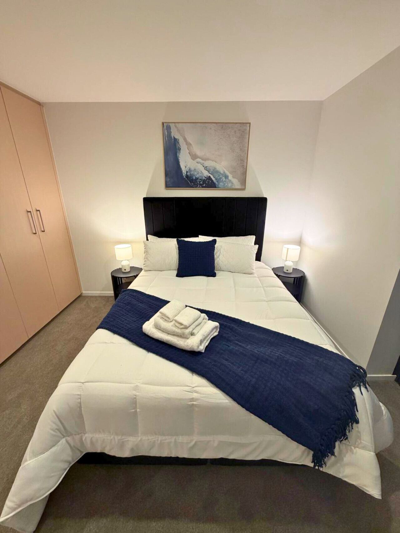 Un lit bien fait, recouvert d’une couverture blanche et bleue, avec deux lampes de chevet et un tableau accroché au-dessus de la tête de lit.