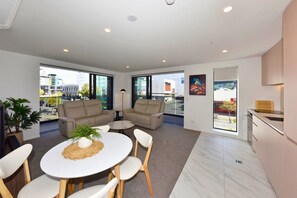 A modern living room with a sofa set, a dining table, and chairs, a small round table with a plant, and a kitchen area with a window. - 2 Bedroom Delight near Te Kaha Stadium (Christchurch)