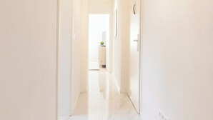 Un pasillo luminoso y blanco con una lámpara en el techo, una puerta a la izquierda y cuadros en la pared derecha.