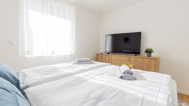 Una habitación de hotel con dos camas, un televisor sobre un mueble de madera y una ventana con cortinas traslúcidas.